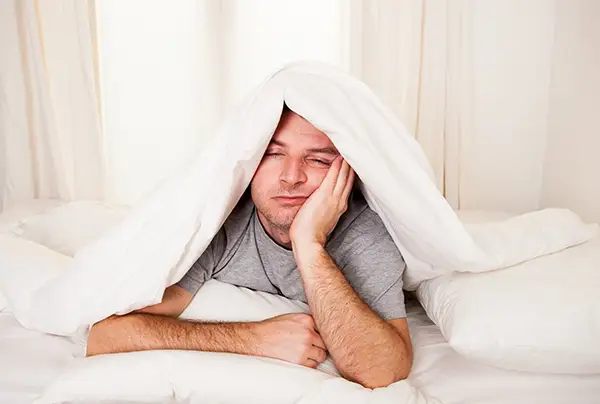 Exhausted man lying under a blanket, looking fatigued due to poor sleep quality caused by sleep apnea.